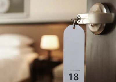 Galerie-Motiv Sonderbau: kleine Hotels und Pensionen. Bild einer offenen Hotelzimmer Tür mit Schlüssel (Nr. 18) und Blick auf ein Doppelbett mit Nachttisch und Lampe.