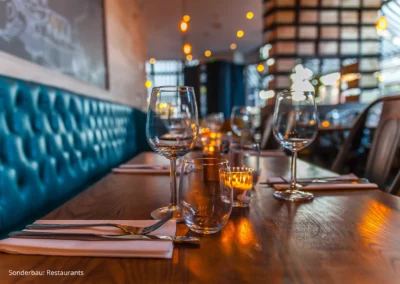 Galerie-Motiv Sonderbau: Restaurants. Bild eines modernen, stylischen Restaurants. Gedekcter Tisch mit Wein- und Wassergläsern, Besteck, Servietten und dunkelblauer Leder-Sitzbank an der Wandseite. Wohlfühl-Ambientebeleuchtung.